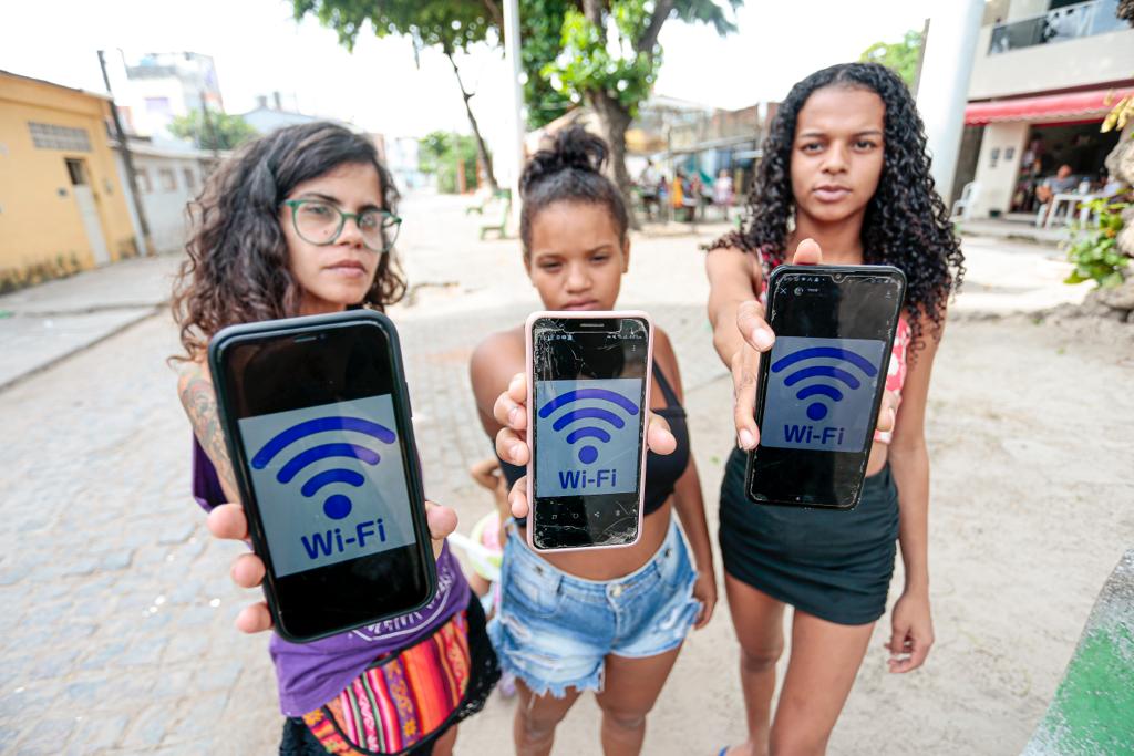 Três mulheres jovens no meio de uma praça, mostram as telas dos seus celulares com o símbolo do wi-fi