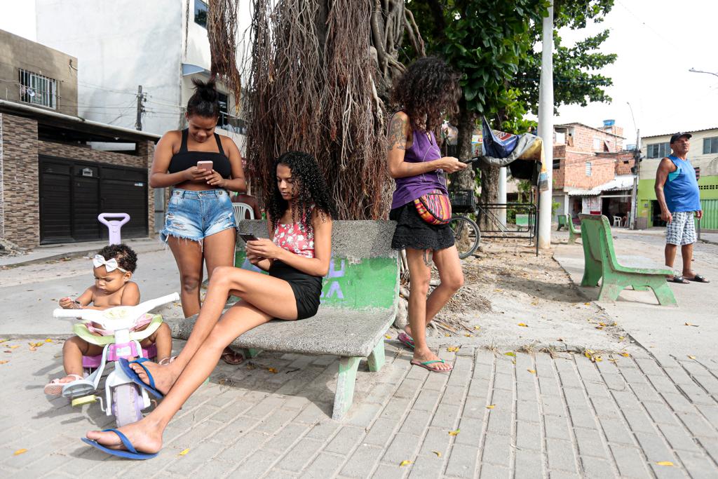 Junto de um carrinho de bebê numa praça pública na periferia do Recife, ao lado de uma árvore de grande porte, três mulheres jovens consultam seus celulares com a cabeça baixa.